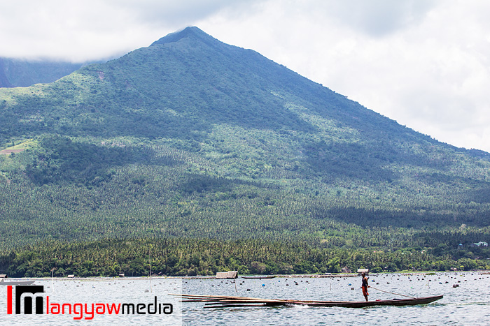 A man stands on his motorized banca as it cruises the placid lake. In the background is the crater of Mt. Asog.